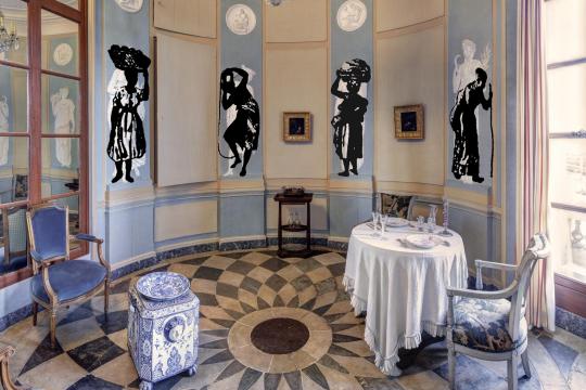Photographie de la salle à manger de la Maison de l'armateur, avec la table dressée. Sur les murs, 4 projections de figures d'esclaves.